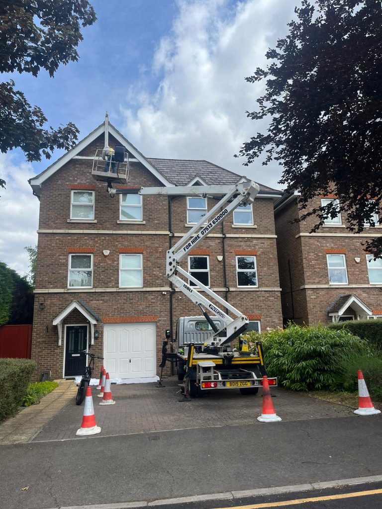 Cherry picker in use at a residential property in Bromley, with a worker elevated near the roof for maintenance or repairs. The vehicle has stabilizing outriggers and is surrounded by safety cones.